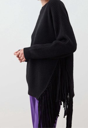 Osoba ubrana w czarny prążkowany sweter z długimi frędzlami po bokach oraz fioletowe spodnie, pokazana z profilu na jednolitym tle.