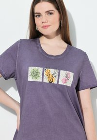 Camiseta de algodón morada con cuello redondo y mangas cortas, con tres cuadrados estampados que muestran gráficos florales y de ciervos.