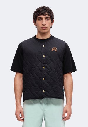 Ung mand iført en sort quiltet vest med guldknapper over en sort t-shirt og lysegrønne bukser, stående mod en ensfarvet baggrund.