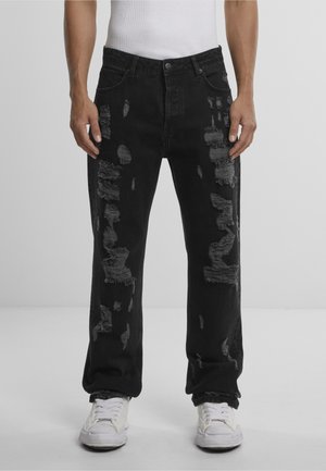 Mees, kes kannab musta kulunud pükse mitme narmendava laiguga ja valgeid tossusid, seisab ühevärvilise heleda tausta ees.