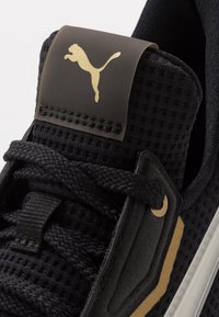 Schwarzer Sportschuh mit strukturiertem Mesh-Obermaterial, goldenem Logo auf einem schwarzen Tab und dicken schwarzen Schnürsenkeln, kombiniert mit einer weißen Außensohle und goldenen Akzenten.
