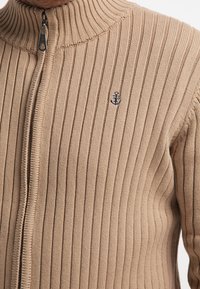 Suéter beige acanalado con un cuello alto y cremallera. Presenta un pequeño logo de ancla en marino en el pecho y un patrón de punto texturizado.