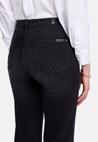 Black denim jeans with a high waist, featuring two back pockets and contrast stitching in a curved pattern. White button-up shirt at the top.