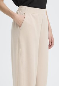Pantaloni beige a gamba larga realizzati in tessuto morbido, con vita elasticizzata e tasche laterali. I dettagli delle cuciture lungo le gambe arricchiscono il design.