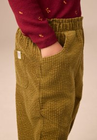 Olijfgroene corduroy broek met verticale ribbels, voorzien van zijzakken en een gathered tailleband, gecombineerd met een kastanjebruine langemouwen shirt.