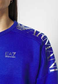 Sweat-shirt bleu avec des accents et des lettrages métalliques, doté d'un col rond et d'un logo sur la poitrine et la manche. Texture douce.