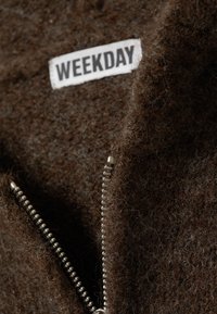 Tissu texturé marron avec une fermeture éclair visible et une étiquette blanche portant la mention "WEEKDAY". Le matériau semble doux et épais.