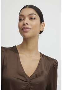 Blusa di raso marrone con scollatura a V, dettaglio arricciato nella parte anteriore e maniche a sbuffo. Tessuto liscio con una leggera lucentezza.