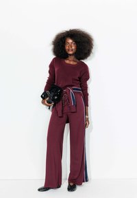 Femme avec un grand afro portant un pull bordeaux et un pantalon large avec des rayures latérales, tenant un sac noir, se tenant devant un mur blanc.