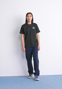 Jonge man met lang donker haar, gekleed in een zwart T-shirt met opdruk, navy broek en blauwe sneakers, staand tegen een witte muur op een blauwe vloer.