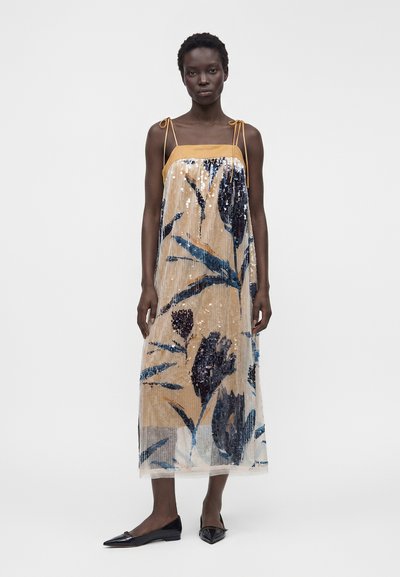 Femme portant une robe midi beige sans manches avec des motifs floraux bleus, des paillettes, des fines bretelles nouées aux épaules et des ballerines noires à bouts pointus.