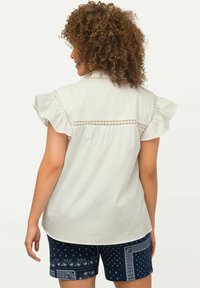 Blouse blanche à manches courtes avec des bords volants et une broderie subtile dans le dos. Associée à un short bleu marine présentant des motifs complexes.