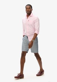 Chemise boutonnière rose clair à manches longues avec une poche poitrine, associée à un short gris et des chaussures bateau bordeaux. Montre en or en accent.
