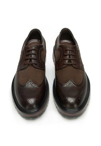 Brązowe buty ze skóry i zamszu z wzorem typu wingtip, zdobieniami brogue oraz grubą czarną podeszwą. Posiadają sznurowadła i gładkie wykończenie.