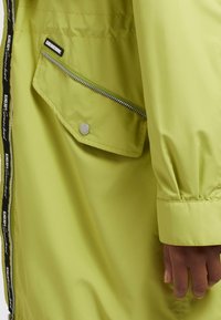Ljusgrön vattentät jacka med en ficka med dragkedja och elastiska ärmslut. Har märkesband längs dragkedjan och en slät, glänsande yta.