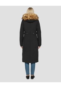 Lange zwarte donzen jas met een capuchon van imitatiebont, voorzien van een getailleerde taille, drukkers aan de achterkant en zijzakken, gedragen over een blauwe spijkerbroek en zwarte schoenen.