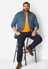 Camicia blu a maniche lunghe sopra una maglietta grafica arancione, pantaloni cargo blu navy e stivali marroni stringati, seduto su uno sgabello.