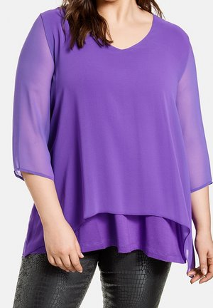 Woman wearing a purple layered V-neck top with sheer three-quarter sleeves and black textured pants, standing with relaxed arms.