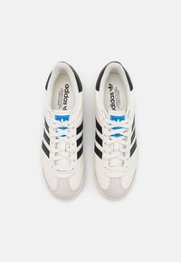 Zapatillas deportivas blancas con rayas y acentos negros, que cuentan con una puntera de ante texturizada, diseño de cordones y un logotipo de marca en azul en la lengüeta.