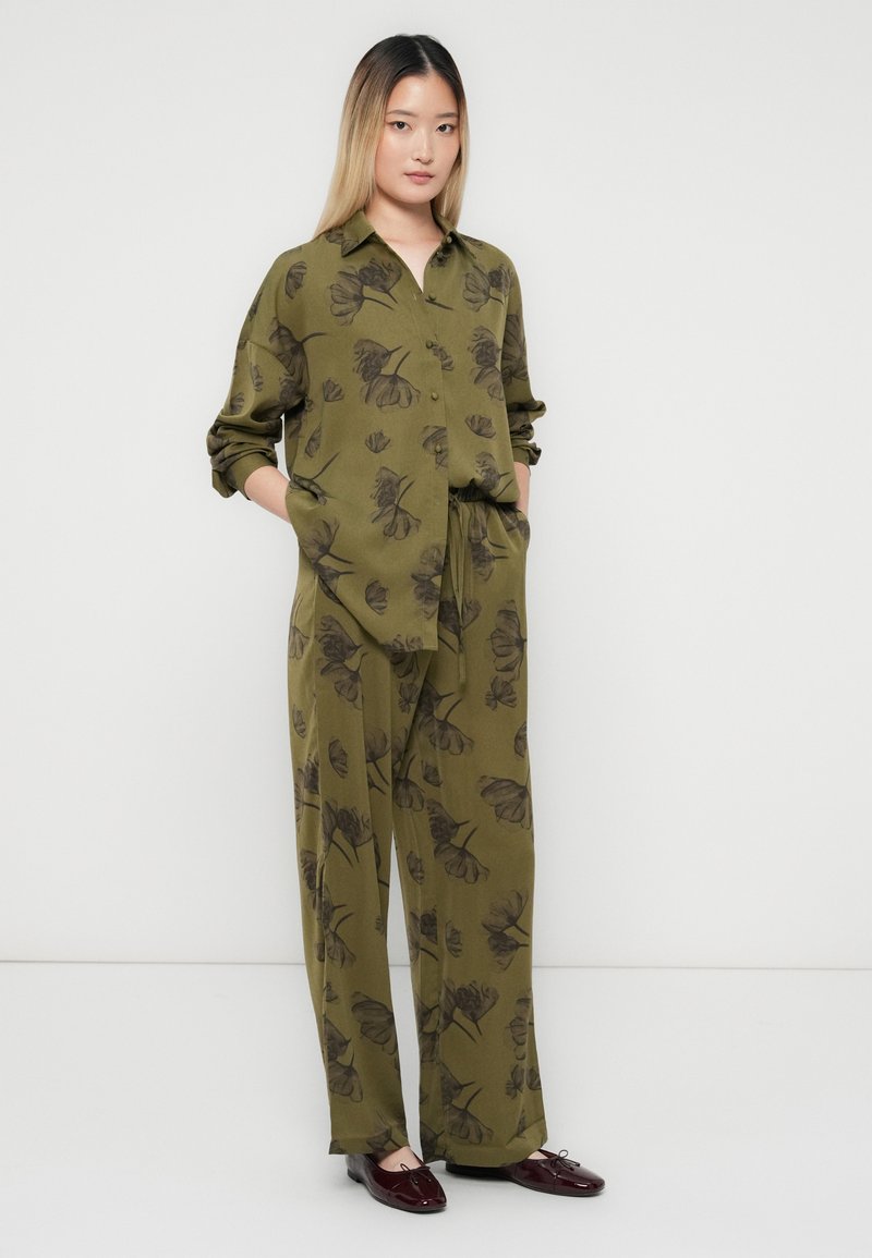 Frau steht mit den Händen in den Taschen, trägt ein olivgrünes, blumiges Hemd mit Knöpfen und passende weite Hosen sowie dunkle burgunderfarbene Schuhe.