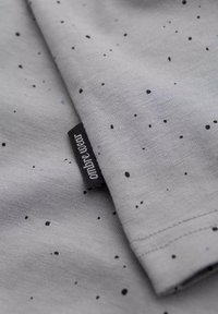 Tejido gris con pequeñas motas negras y azules, con una etiqueta de marca negra y texto blanco. Textura suave y diseño casual.