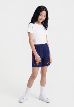 Dziewczyna stojąca, ubrana w białą koszulkę, granatowe spodenki piłkarskie FC Barcelona z logo, białe skarpetki i białe trampki, uśmiechająca się i patrząca w dół.