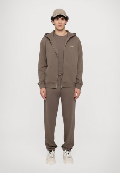 Hanorac maro cu fermoar frontal, pantaloni de trening asortati si o sapca. Tesatura pare moale, cu un design minimal si detalii de logo.