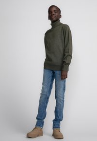 Maglione a dolcevita verde oliva, design aderente. Jeans slim azzurri chiari. Stivaletti beige con suola testurizzata. Estetica semplice e moderna.