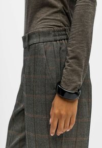 Bracelet noir brillant sur une main, associé à un pantalon à motif pied de poule gris avec un léger carreau rouge, et un haut à manches longues marron.