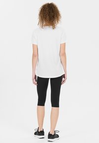 Vit kortärmad träningströja med rundad nederkant, ihopparad med svarta capribyxor och svarta träningstränare. Lockigt hår synligt i nacken.