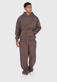 Giovane uomo che indossa un felpa oversize marrone e pantaloni della tuta abbinati, con sneakers bianche, in piedi con una mano in tasca.