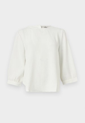 Blusa bianca a maniche lunghe con scollo rotondo, tessuto leggermente strutturato e polsini arricciati su uno sfondo chiaro e uniforme.