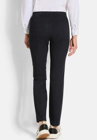 Vrouw in zwarte slim-fit broek en beige sneakers met zwarte accenten, staande tegen een effen lichte achtergrond, van achteren gezien.