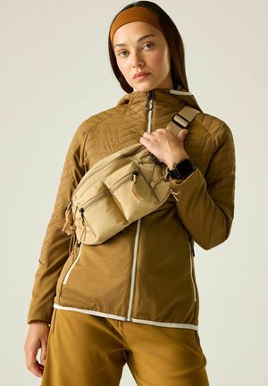 Junge Frau trägt braune Outdoorjacke und Stirnband, hat eine beige Umhängetasche und eine Smartwatch am Handgelenk, steht vor einem einfarbigen Hintergrund.