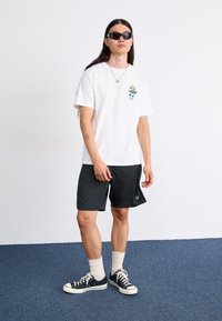 Hvid bomulds T-shirt med et farverigt grafisk design, parret med sorte shorts og mørke sneakers. Modellen bærer solbriller og ankelstrømper.