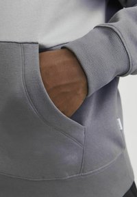 Grauer Pullover mit geripptem Bündchen und Kängurutasche. Weicher Stoff mit glatter Textur. Arm ist teilweise in der Tasche sichtbar.