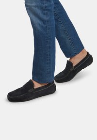 Svarta mockaloafers med rund tå och sydda detaljer, bärs med blå denimjeans, vilket visar en avslappnad och bekväm design.