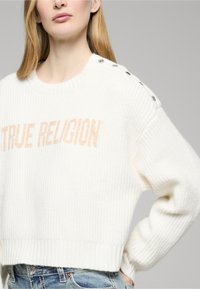 Maglione bianco a maglia con texture a coste, con logo "TRUE RELIGION" in beige chiaro e bottoni decorativi sulla spalla sinistra.