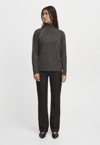 Temno siva valovita turtleneck pulover s polovičnim zadrgo, v kombinaciji z črnimi hlačami ravnega kroja in čevlji s koničastim nosom.