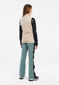 Beige Fleece-Weste mit hohem Kragen, getragen über einem langärmeligen schwarzen Hemd mit weißen Sternmustern und türkisfarbenen Hosen mit schwarzen Seitenstreifen.