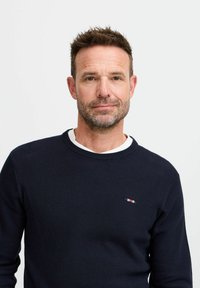 Mørkeblå strikkegenser med teksturert overflate og rund hals. Har en liten rød, hvit og blå logo på brystet.