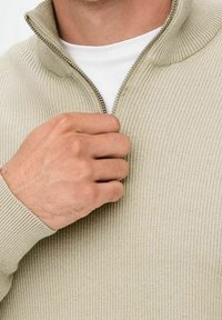 Beige ribstrikket sweater med lynlåsdetaill ved halsen. Close-up af en hånd, der griber fat i lynlåsen. Hvid undertrøje synlig under sweateren.