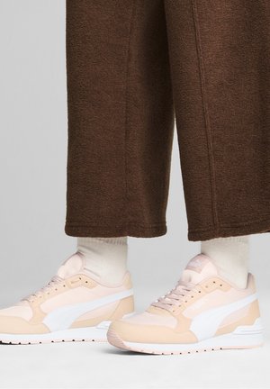 Pieds portant des baskets Puma rose clair et beige avec un logo blanc, des chaussettes crème et un pantalon marron à jambes larges.