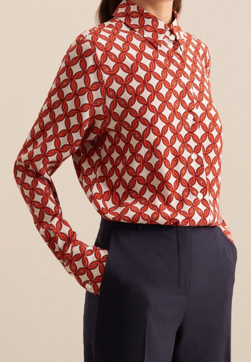 Kvinna i en långärmad skjorta med knappar i ett röd kedjemönster, instoppad i mörknavybyxor.