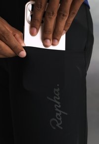 Hand steckt ein weißes Smartphone mit Dual-Kameras in die schwarze Hosentasche mit „Rapha“-Logo.