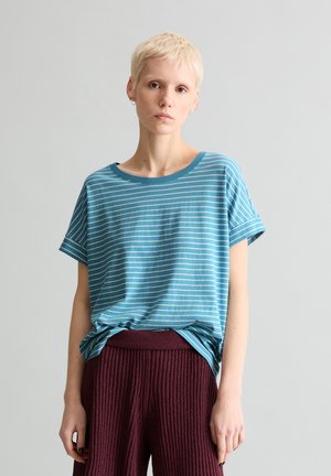 Persoon met kort blond haar, gekleed in een blauw-wit gestreept T-shirt, ingestopt in een textuurrijke bordeauxrode broek, staand tegen een effen lichte achtergrond.