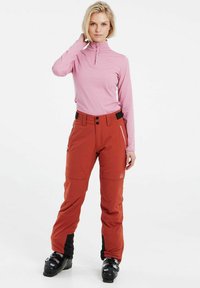 Light pink, long-sleeve top with a zip neck; orange snow pants with pockets and black accents; black ski boots.