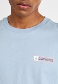 Lysblå bomuldstrøje med rund hals. Har et rødt og hvidt rektangulært logo med teksten "CONVERSE" og et stjernemblem.