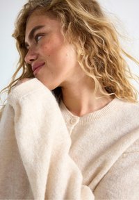 Beige strikket cardigan med rund halsudskæring, knapdetalje og lange, løse ærmer lavet af et blødt, tekstureret garn.