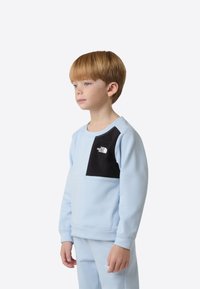 Sudadera azul claro con parche negro, cuello redondo y puños acanalados. Presenta un logo en el pecho. Textura suave, ajuste estructurado.